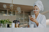 Photo of attractive young woman wears bath towel on head and soft white dressing gown holds mobile phone enjoys online communication applies moisturising undereye patches drinks hot beverage