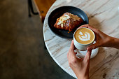 Over the shoulder view of an Asian woman enjoying her breakfast, a croissant with a cup of coffee in a cafe during the morning