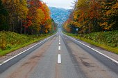 A diminishing perspective view of a country highway running straight uphill toward a mountain in background with vibrant fall colors by the road on a sunny autumn day in Kamishihoro, Hokkaido, Japan