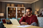 A busy young adult male freelancer resting his head on his hand while working on a project on a laptop.