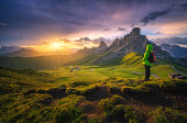 Woman with backpack on the trail and beautiful alpine mountain peaks at colorful sunset in summer. Passo Giau, Dolomites, Italy. View of girl, rocks, hills, green grass, flowers, vibrant sky. Hiking