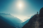 Standing woman on the hill against mountain valley at bright sunny day. Landscape with girl, trail, mountain, blue sky with sun and low clouds at sunset in Nepal.  Lifestyle, travel. Trekking